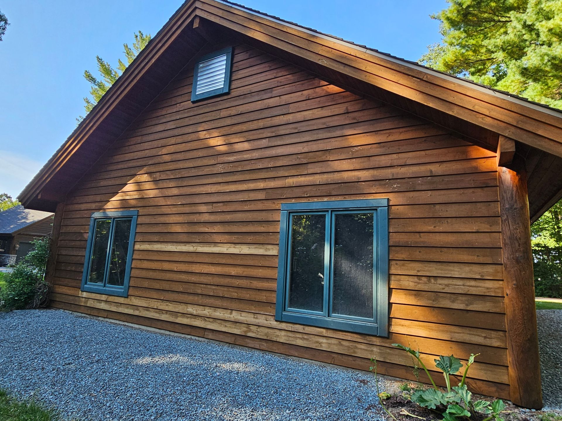 Brown wooden cabin with green-framed windows, against a blue sky. Gravel and greenery in the foreground.
