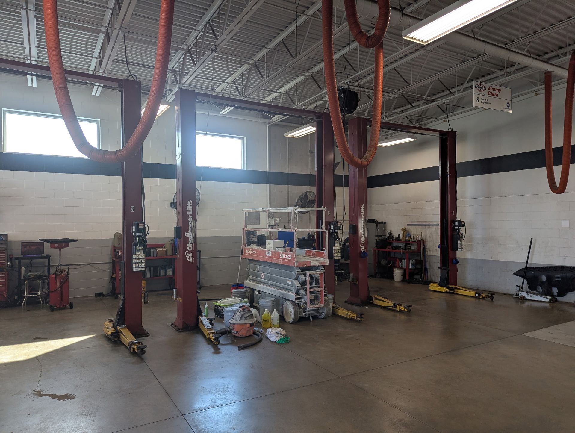 Inside a car repair shop: red car lifts, equipment, concrete floor, white walls, and bright overhead lights.