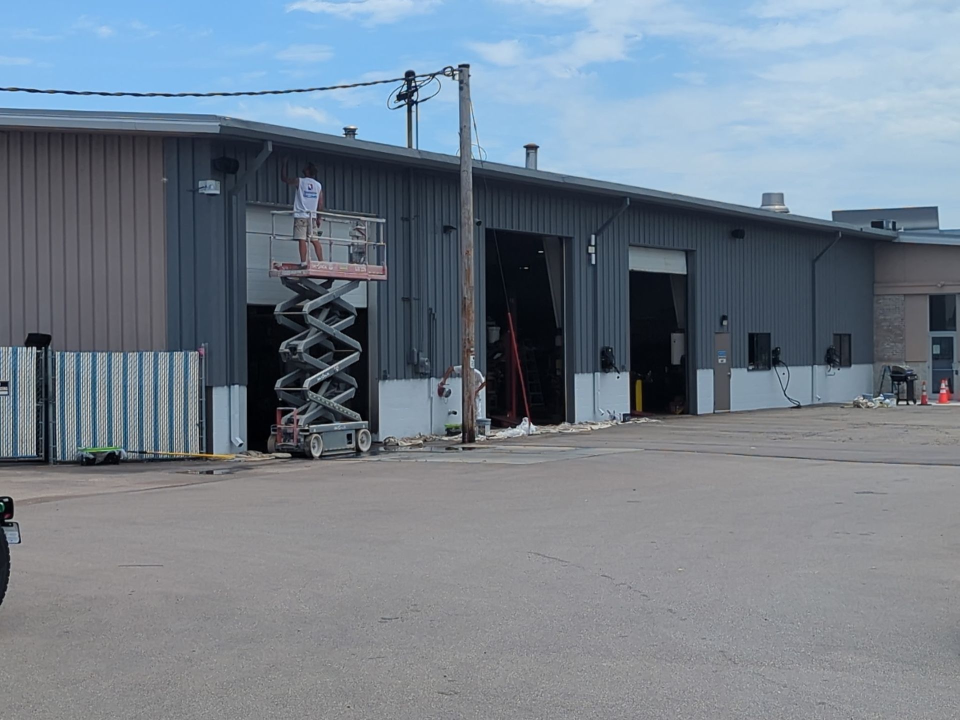 Person on a scissor lift near a gray industrial building. Overhead power lines and open garage doors are visible.