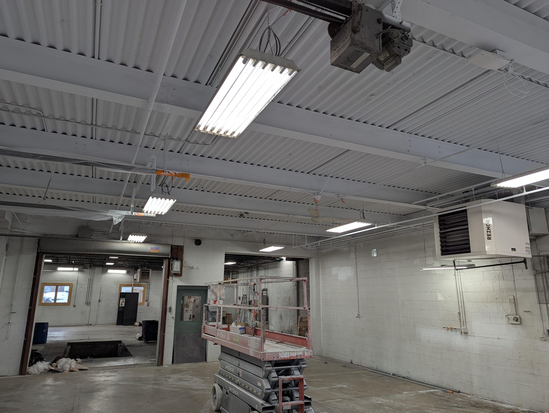 Interior of a commercial space with corrugated metal ceiling, fluorescent lights, and lift, under renovation.