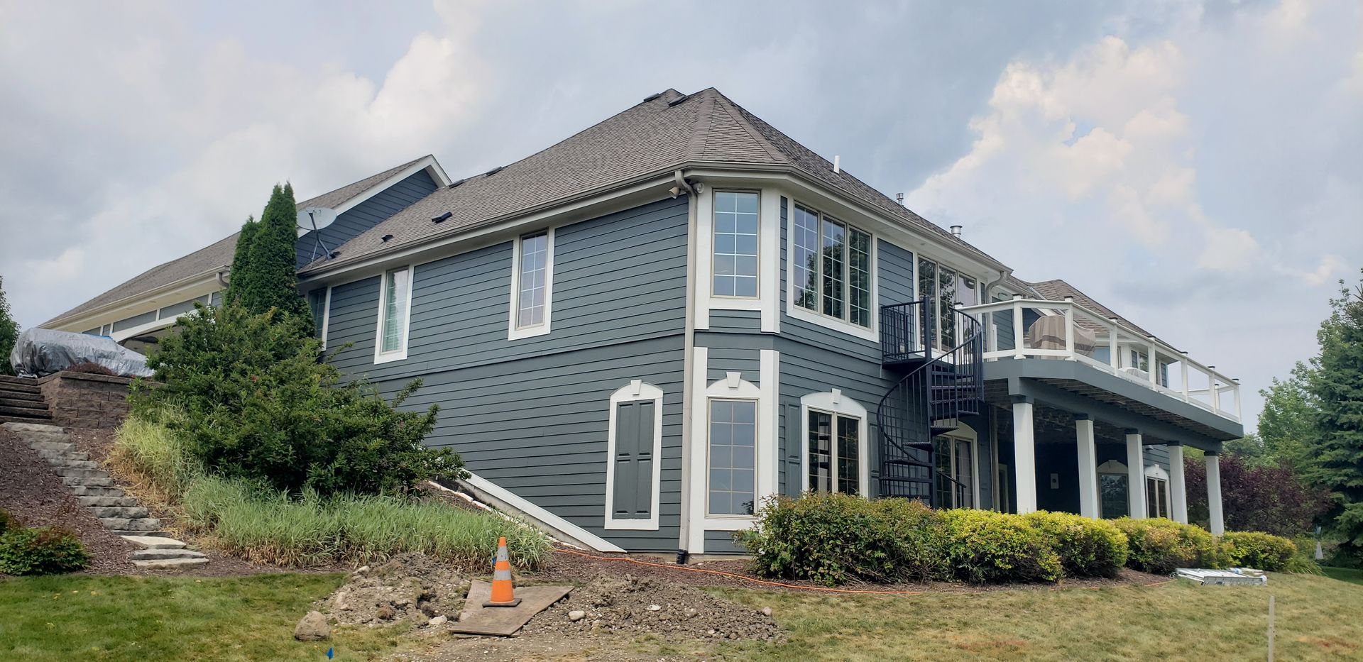 Large gray house with white trim, set on a green grassy hill with landscaping, under a cloudy sky.