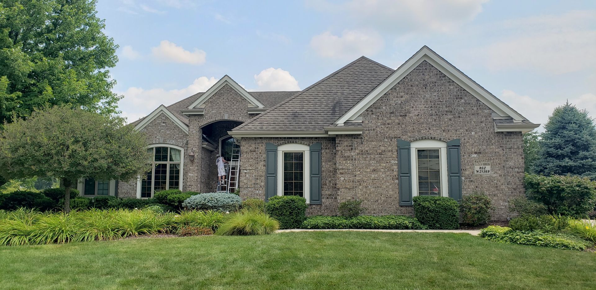 Brick house with gray roof, shutters, and landscaping. Green lawn and a bright sky are visible.
