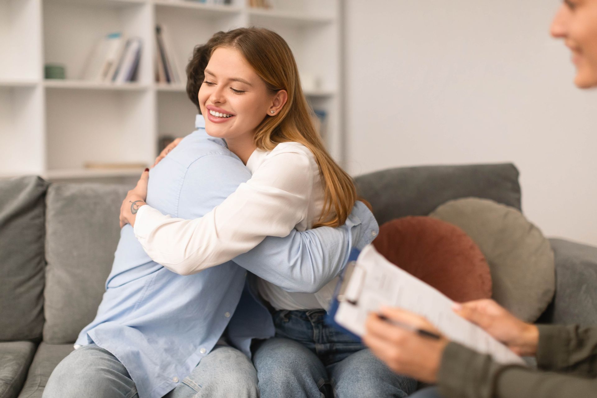 Couple embraces on a couch, smiling at each other, as a therapist looks on.
