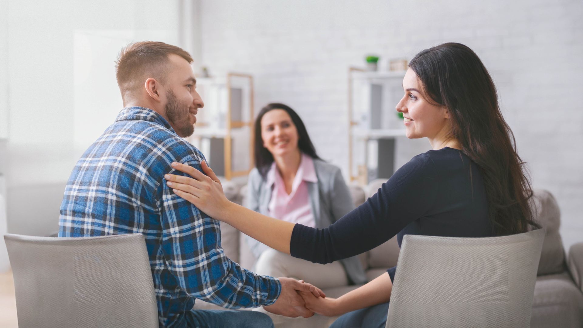 Couple holding hands and smiling at each other, with a therapist smiling in the background, in a counseling session.