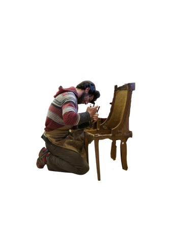 A man kneels, working on a wooden chair. He wears a red and white striped sweater.