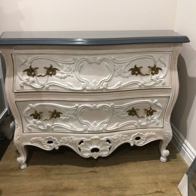 White ornate two-drawer chest with gold hardware and gray top, on a wood floor.
