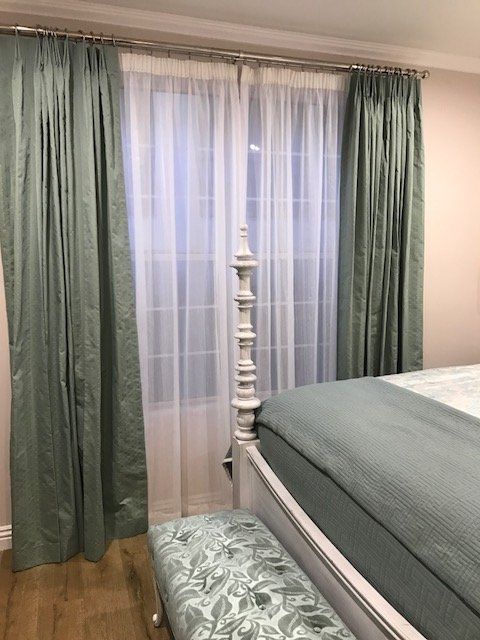 Bedroom with sage green curtains, sheer white curtains, white bed frame, and patterned bench.