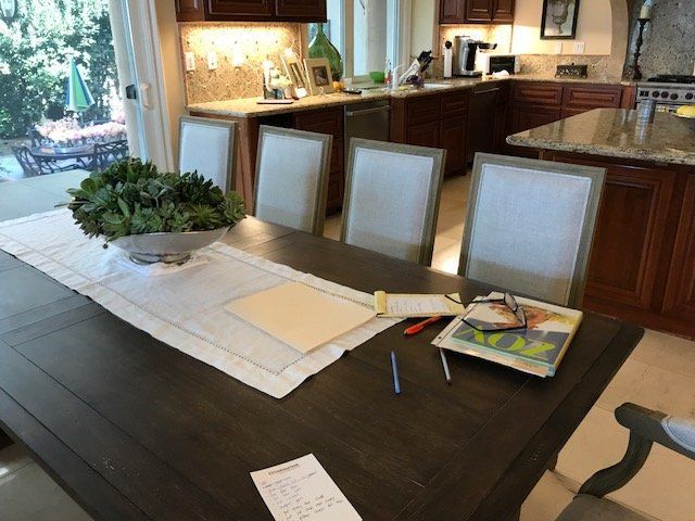 Dining table with four chairs, set in front of a kitchen. Table has documents, a plant, and a placemat.