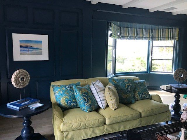 Living room with blue panel walls, green couch, turquoise pillows, and bay window with striped shade.