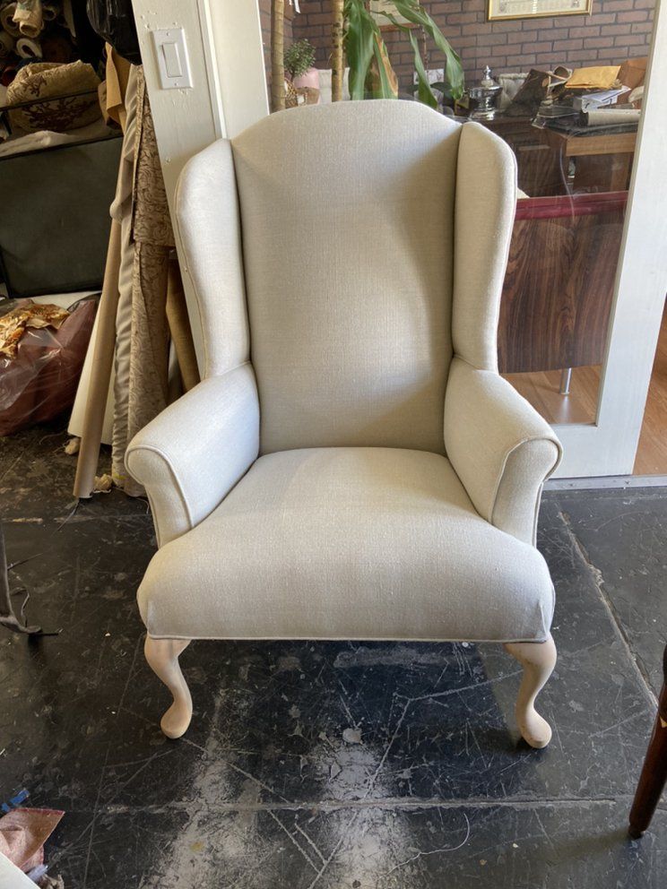 Cream-colored upholstered wingback chair with light wood legs on a dark floor in a workshop setting.