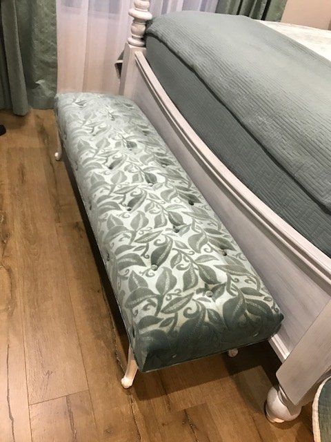 Tufted bench with leafy print at the foot of a bed with a light blue cover, on a wood floor.