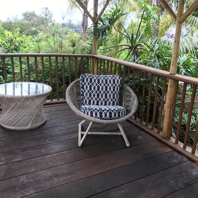 Deck with wicker chair, glass-top table, and railing overlooking lush green plants.