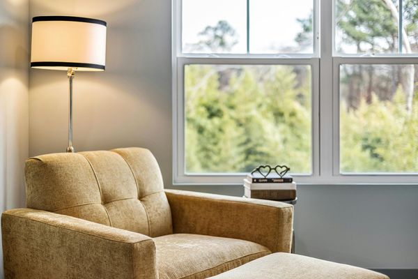 Beige armchair with a matching footstool next to a window overlooking trees. A lamp is next to the chair.