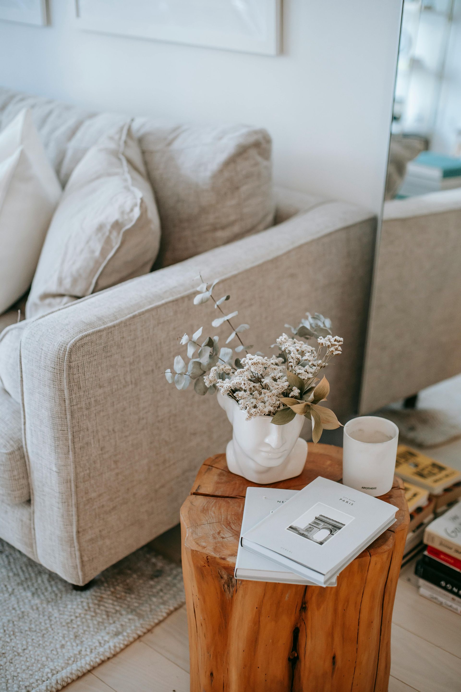 Cozy living room: beige couch, wooden side table with floral arrangement and books, neutral colors.