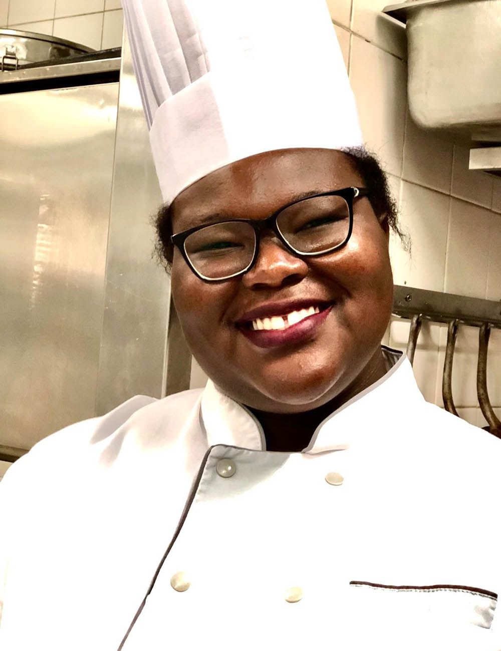 Un chef souriant, portant toque et veste de chef, dans une cuisine.