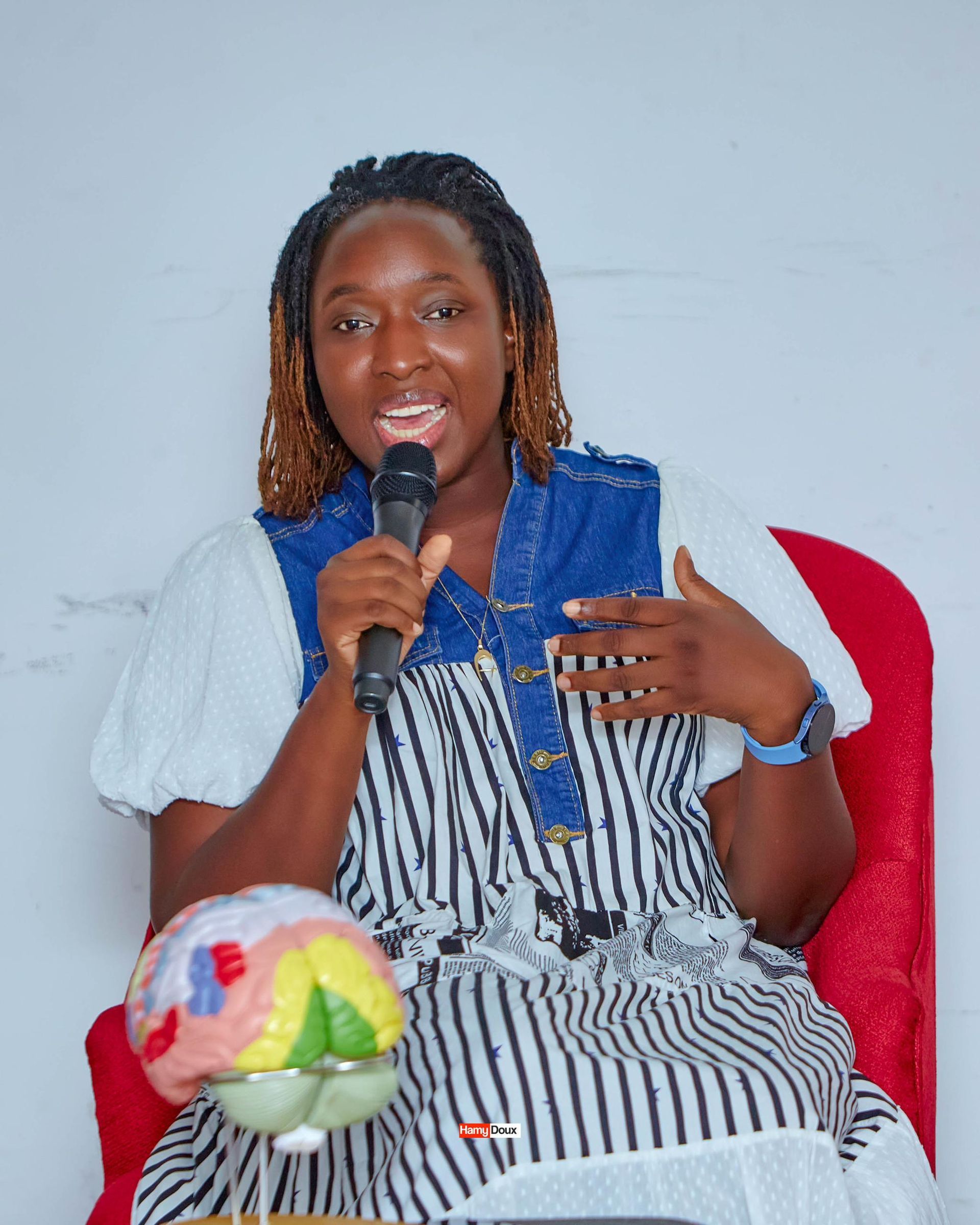 Une personne aux cheveux tressés tient un microphone et gesticule, assise sur une chaise rouge