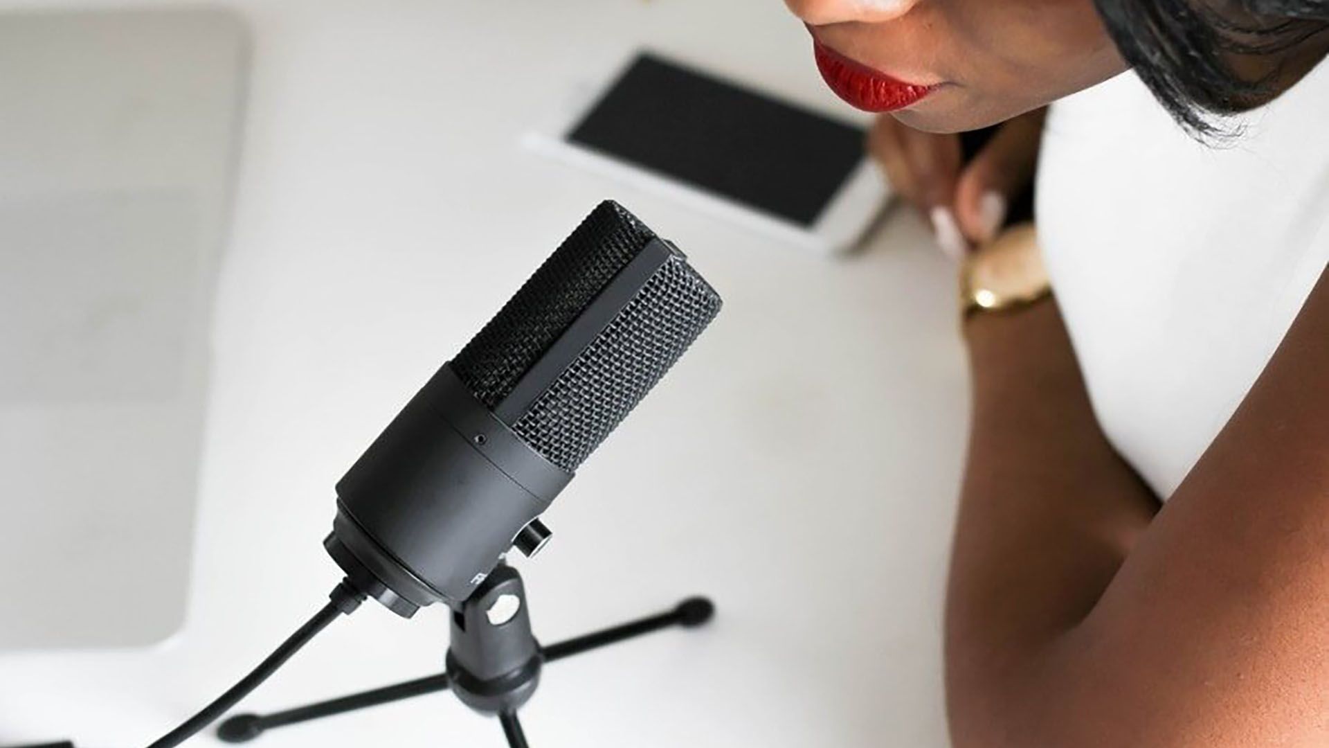 Une personne au rouge à lèvres rouge parle dans un microphone noir sur pied. Bureau blanc, téléphone, montre.