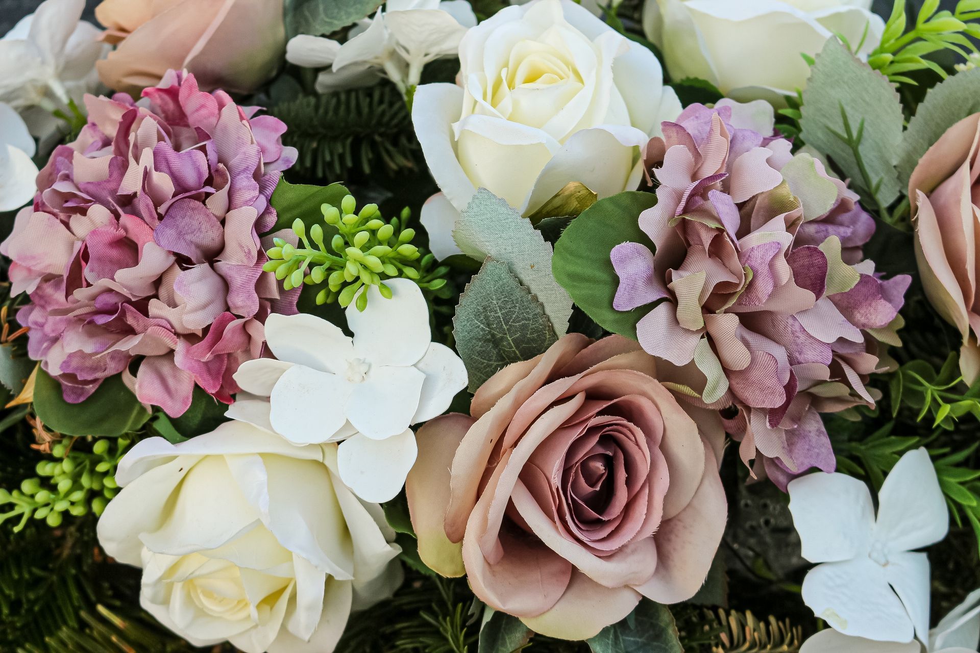 A close-up of an artificial floral arrangement featuring mauve roses, white roses, and purple hydrangea blossoms.