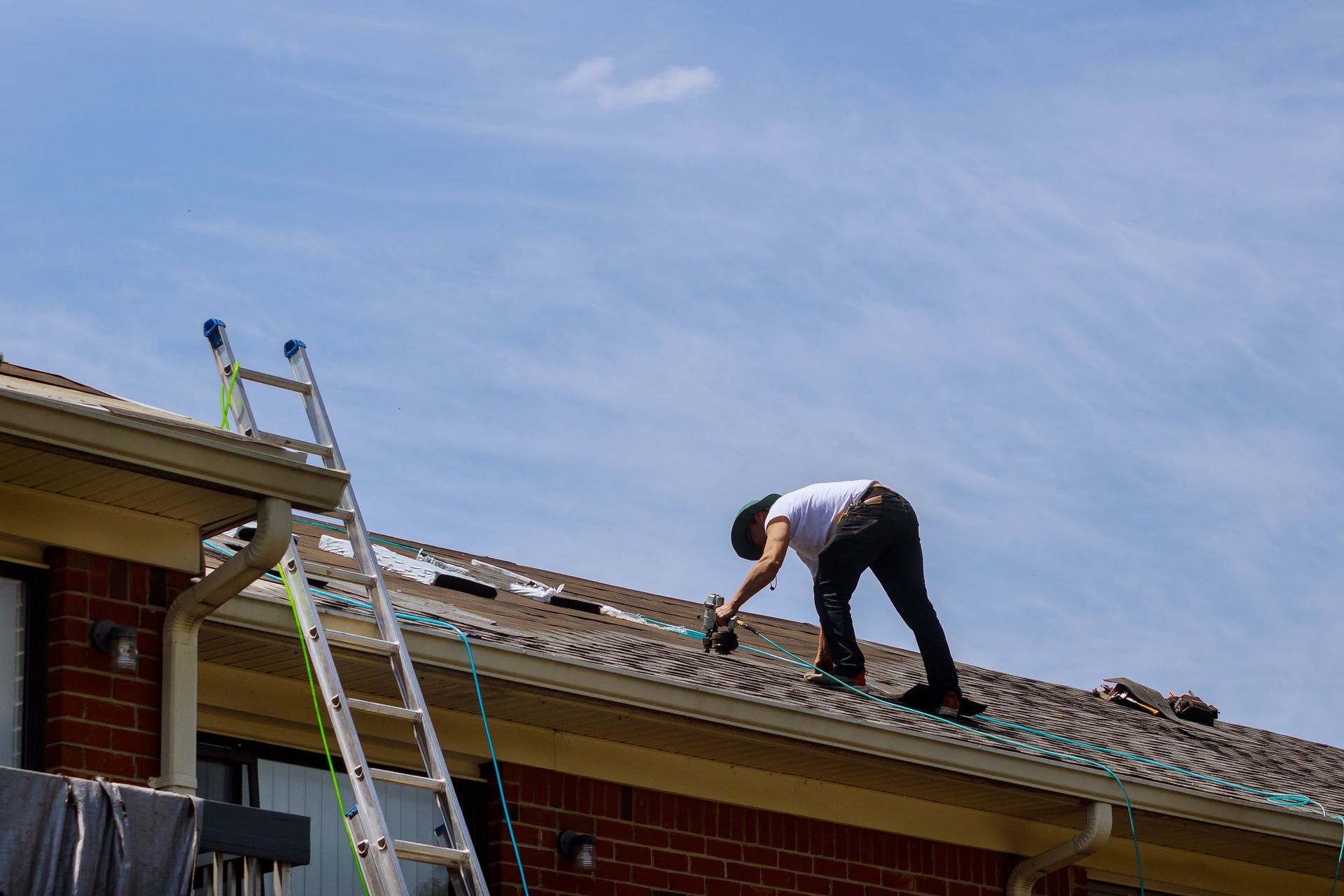 Worker on a roof repairing shingles beside a ladder on a brick house