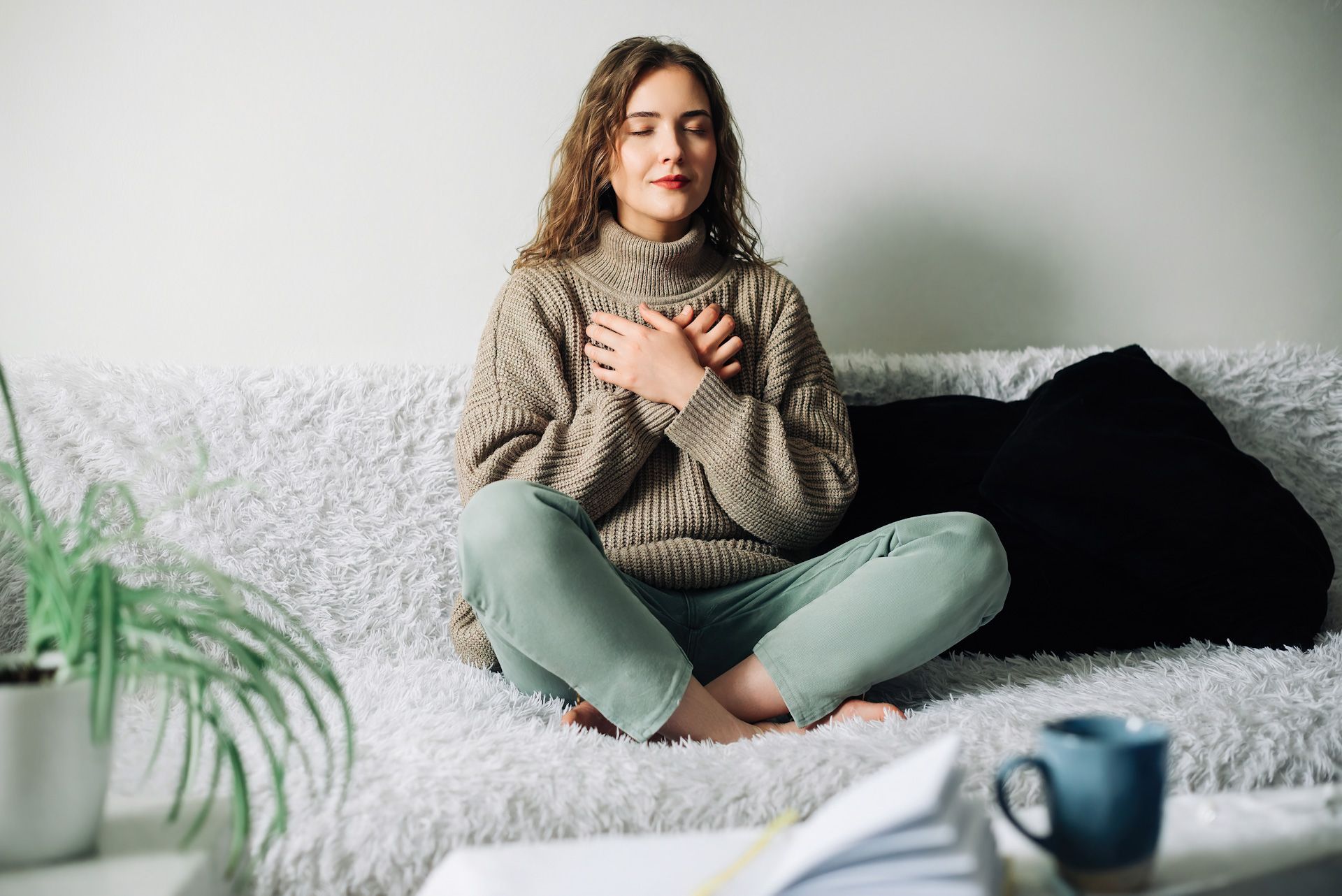 Woman sitting cross-legged on a couch with hands on chest, eyes closed, appearing to meditate.