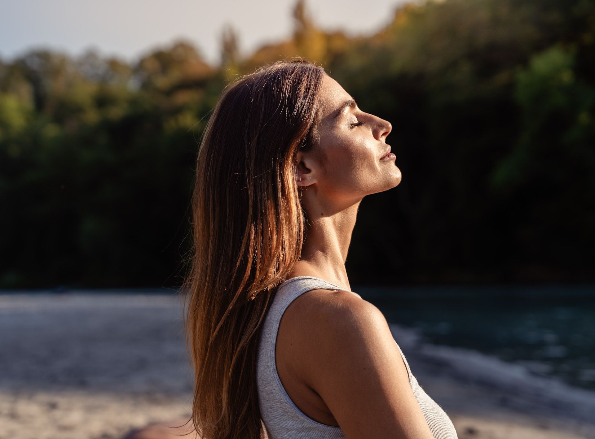 Woman with closed eyes, head tilted up, taking a deep breath outdoors with sunlight on her face.