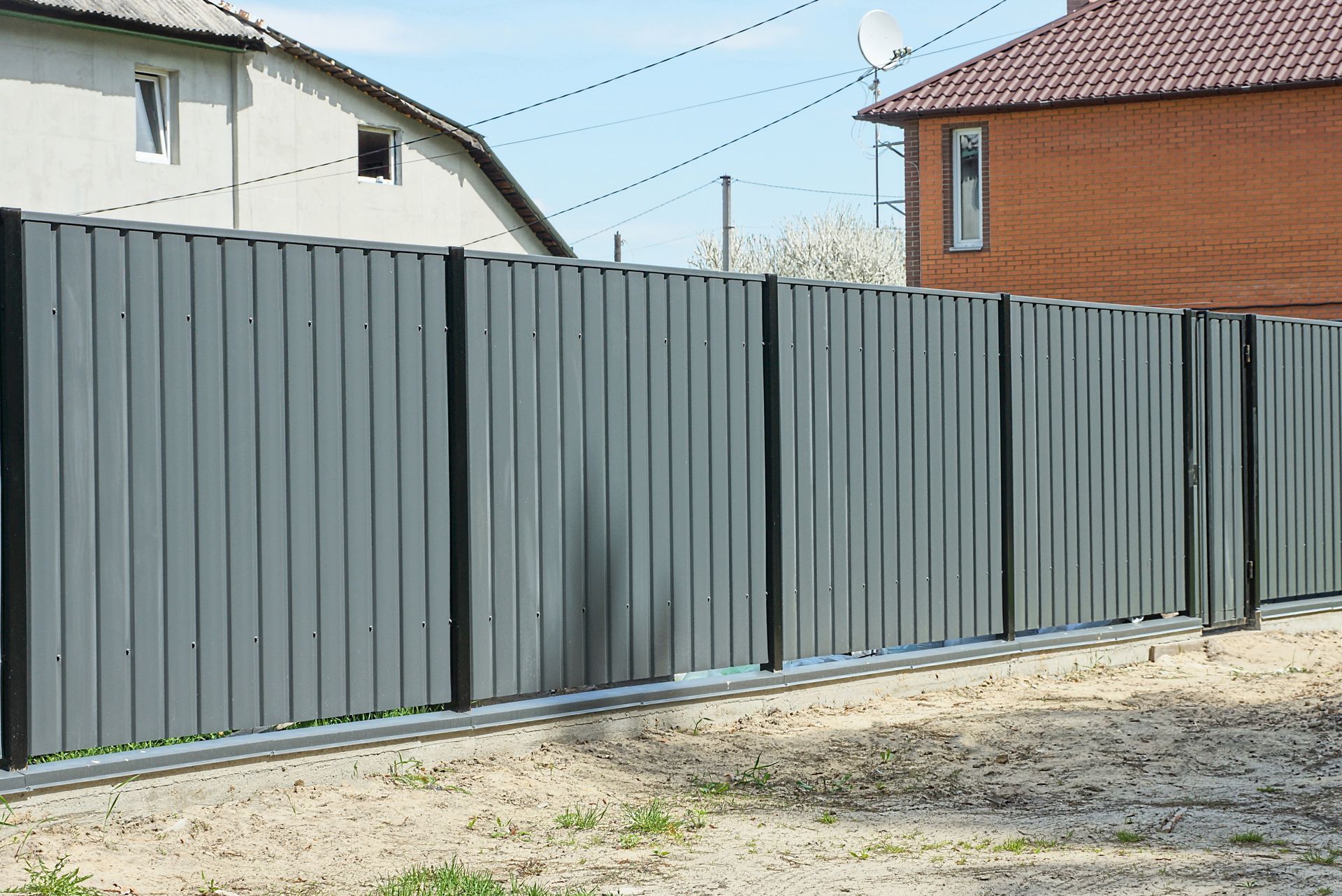 Gray metal fence with vertical lines, separating a sandy yard from houses.