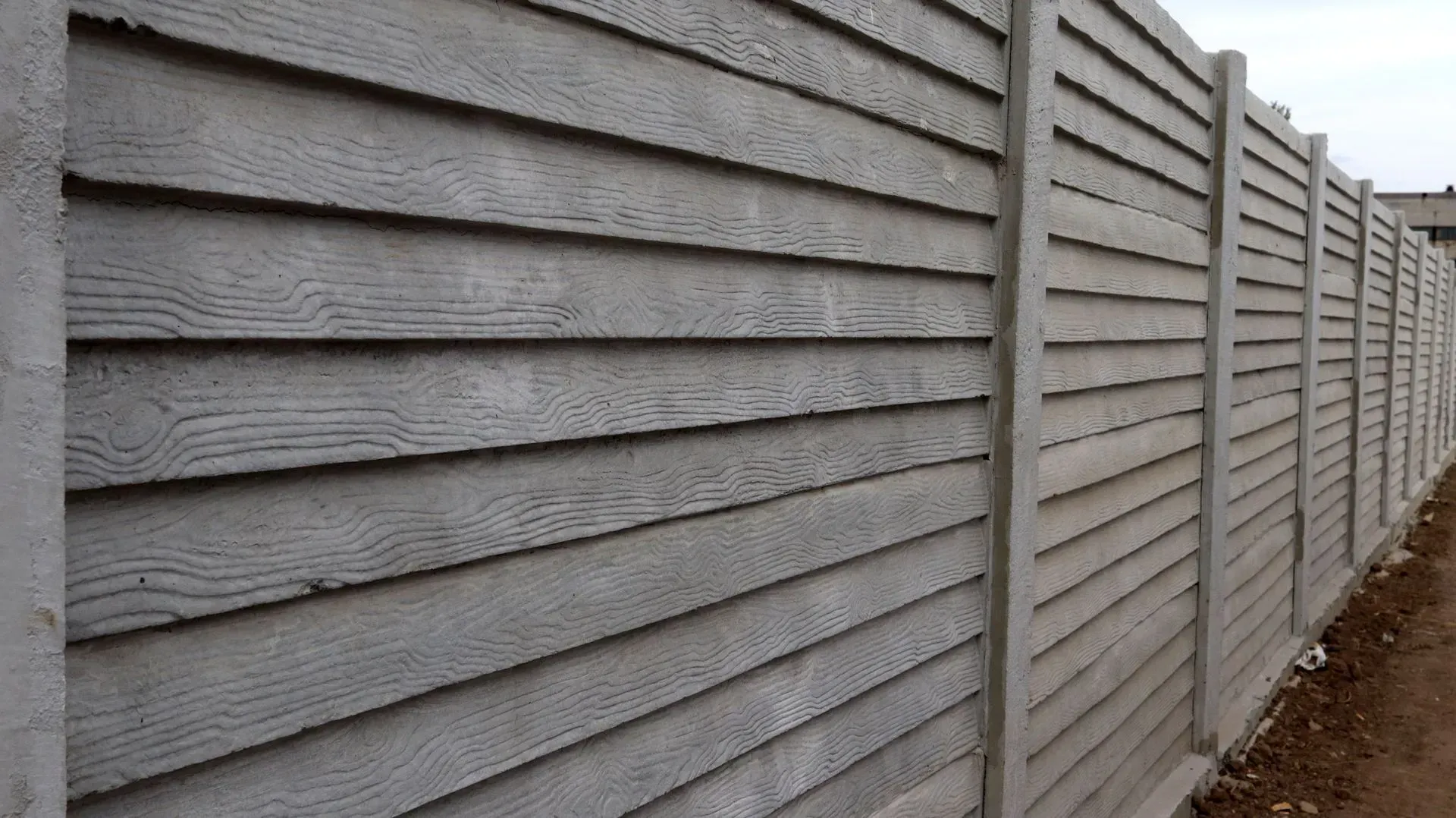 Gray concrete fence with horizontal wood-like paneling, running along an outdoor path.