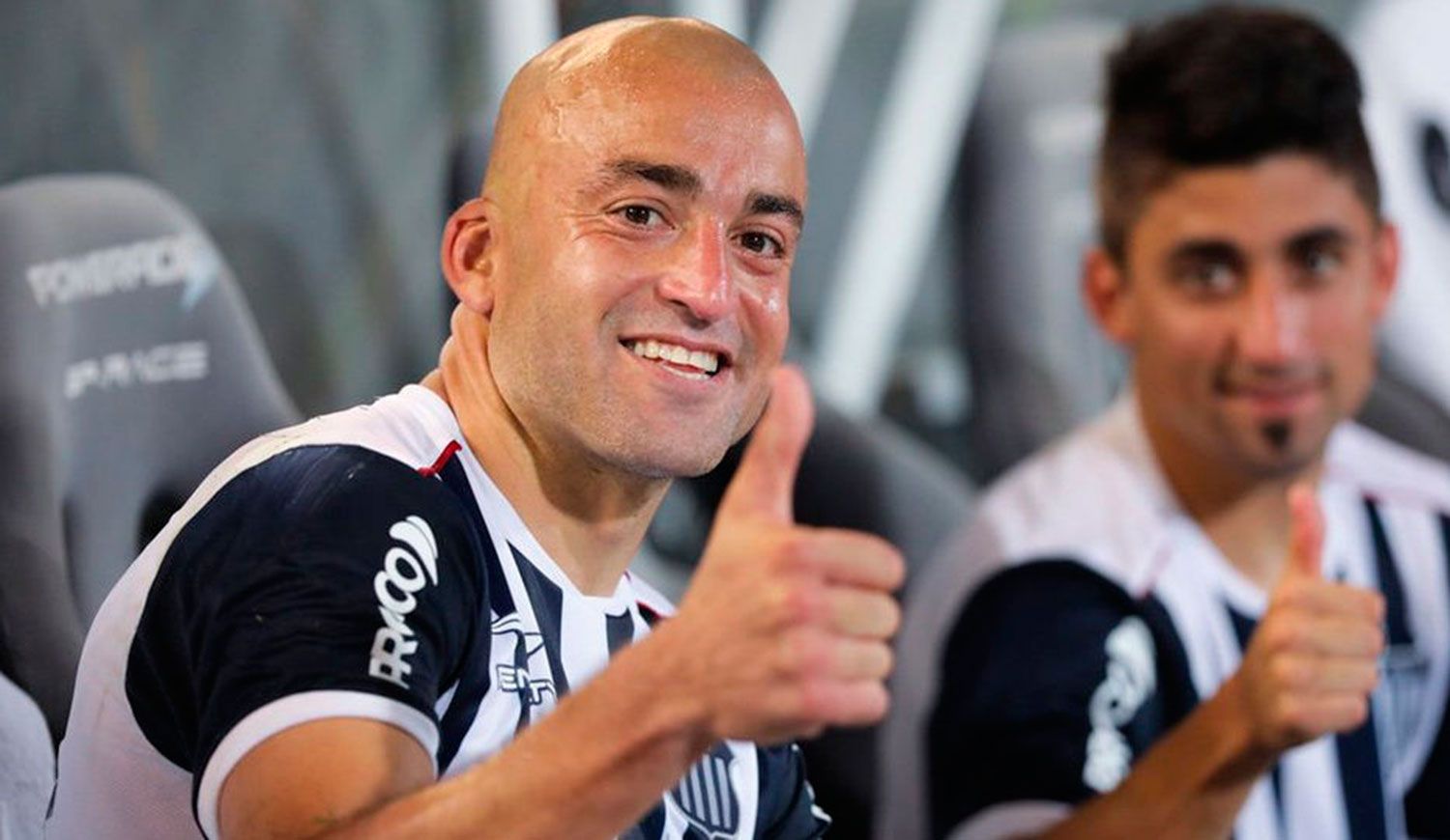 Jugador de fútbol haciendo un gesto de pulgar hacia arriba, sentado en un banco, vistiendo una camiseta blanca y negra.