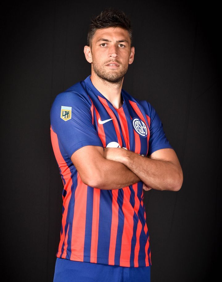 Jugador de fútbol con una camiseta de rayas azules y rojas, con los brazos cruzados, de pie sobre un fondo oscuro.