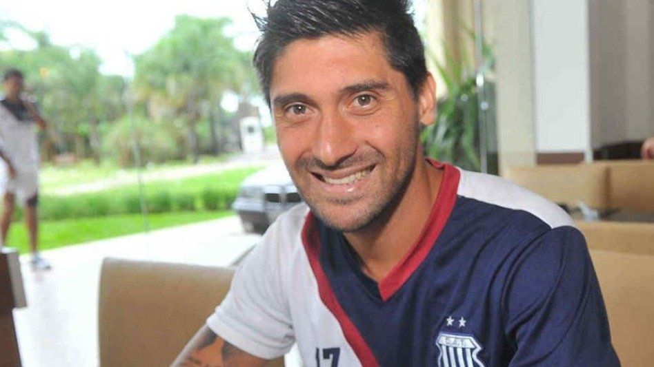 Un hombre con una camiseta de fútbol azul marino y blanca sonríe, sentado en una mesa.