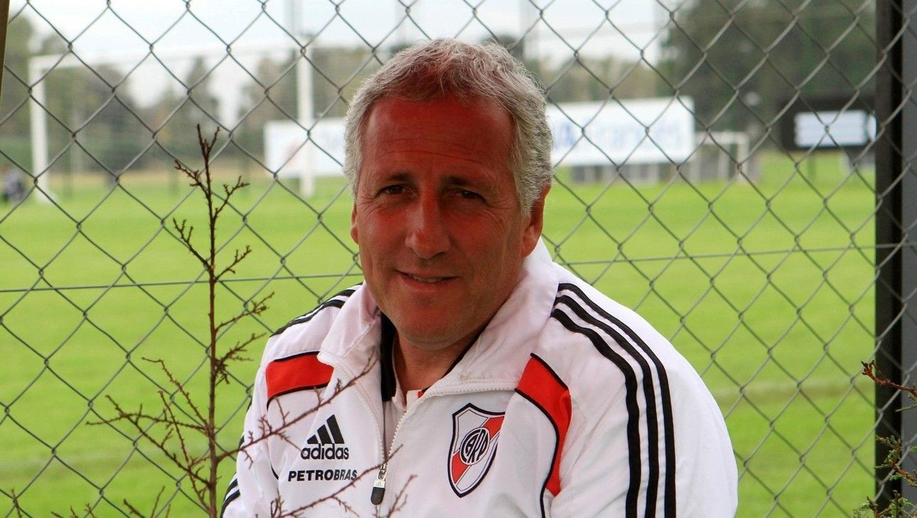 Hombre con chaqueta deportiva blanca sonriendo frente a una valla de alambre y un campo de fútbol.