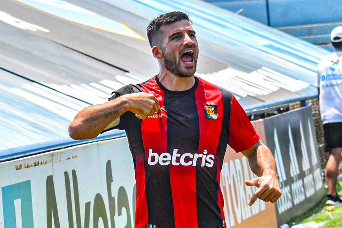 Jugador de fútbol con camiseta roja y negra celebrando con los brazos levantados en un campo.