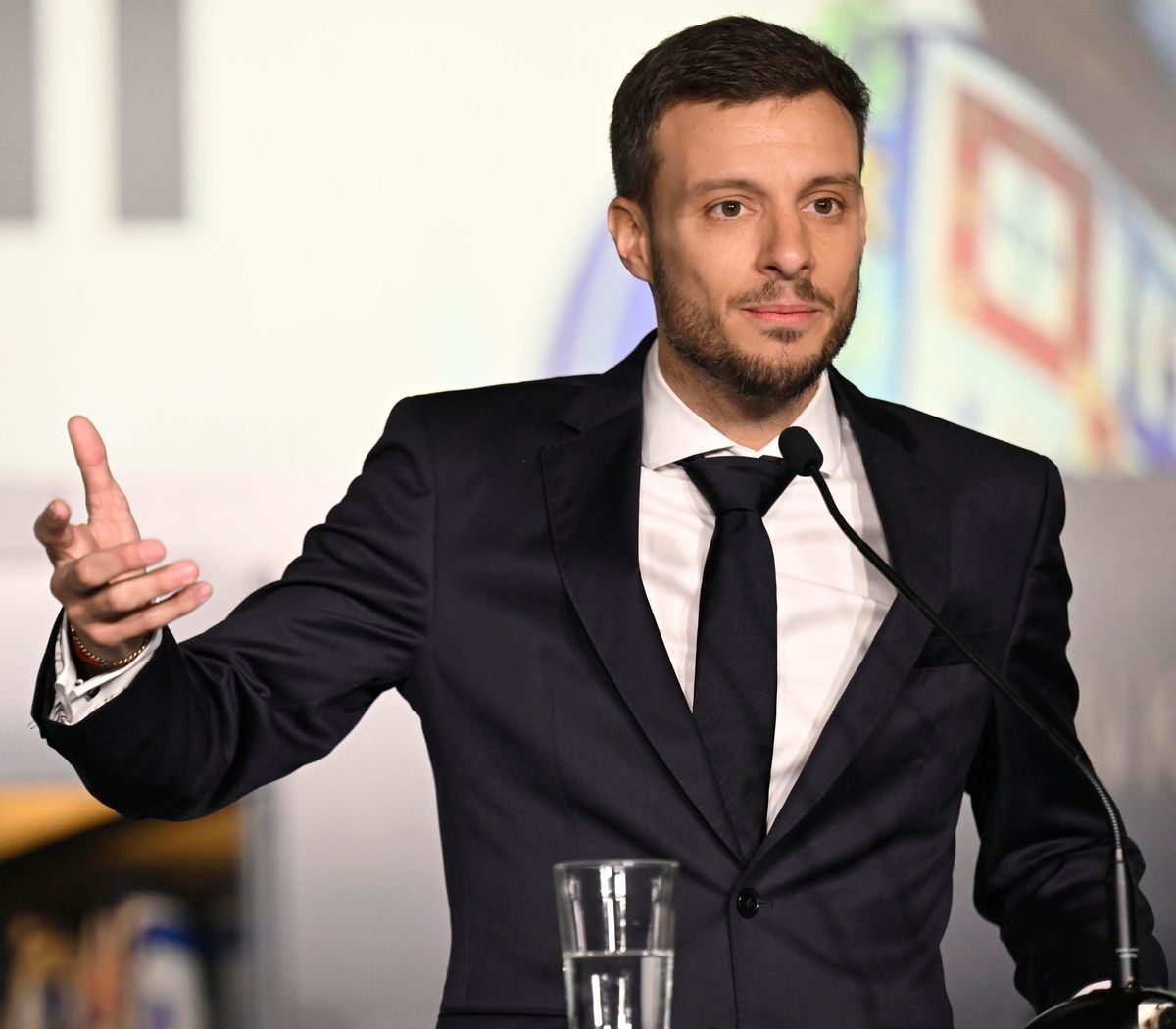 Un hombre con traje oscuro habla en un podio, gesticulando con la mano izquierda. Tiene expresión seria.