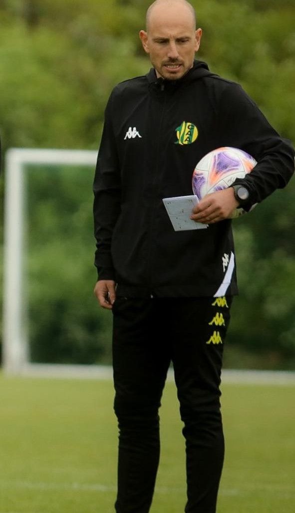 Hombre con chándal negro sosteniendo un balón de fútbol y una tableta en un campo, cerca de una portería.