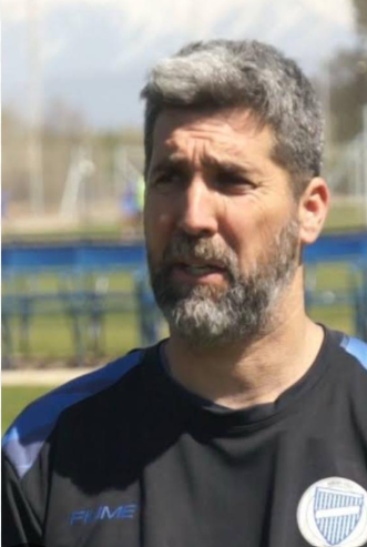 Entrenador de fútbol desde la banda, señalando y dando instrucciones. Vestía camiseta y pantalones negros, y reloj.