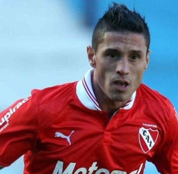 Jugador de fútbol con camiseta roja, pelo corto oscuro, mirando hacia adelante, parche con