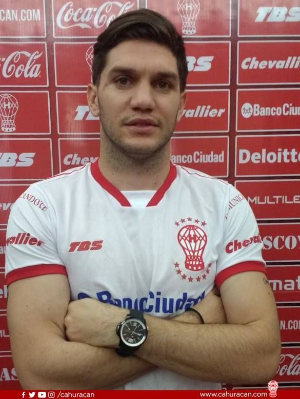 Jugador de fútbol con camiseta blanca y los brazos cruzados, delante de un fondo promocional rojo y blanco.