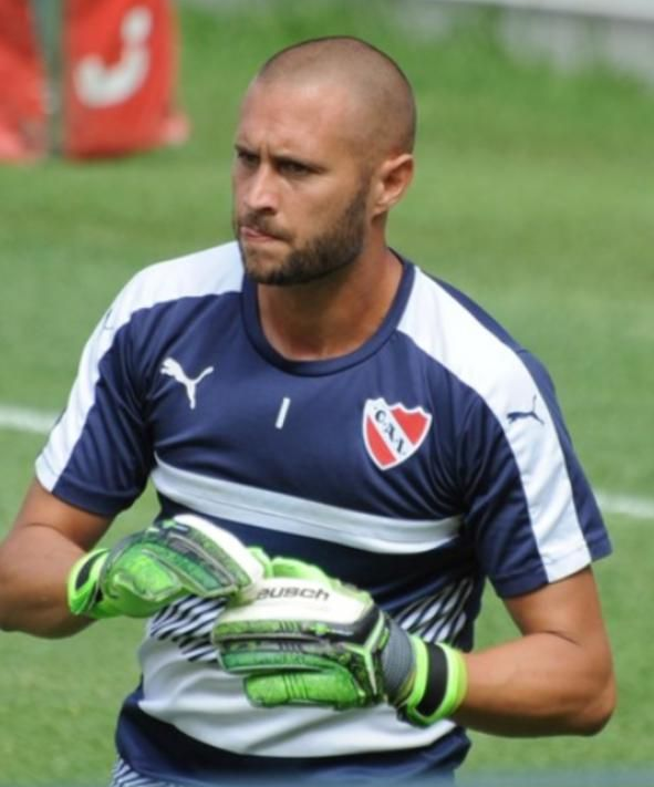 Portero de fútbol con camiseta azul y guantes verdes, mirando concentrado con la cabeza rapada.