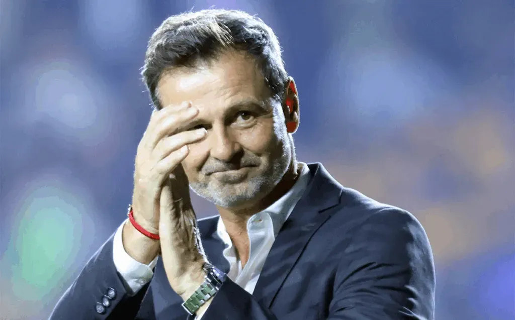 Hombre de pelo gris, vestido con una chaqueta azul marino, aplaudiendo y con aspecto pensativo.