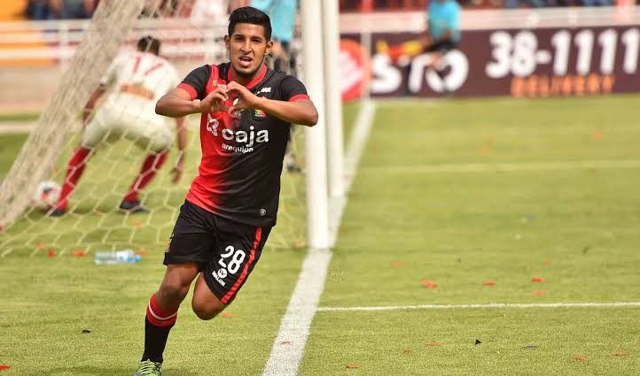 Jugador de fútbol con camiseta negra y roja celebrando, formando un corazón con sus manos cerca de la portería.