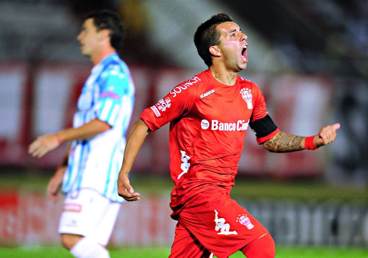 Jugador de fútbol con uniforme rojo celebrando con la boca abierta y el brazo en alto. Otro jugador de azul detrás de él.