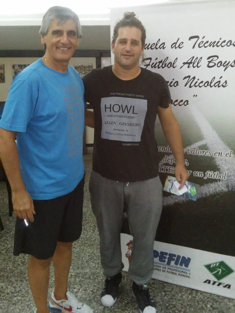 Dos hombres posan juntos; uno con camisa azul y pantalones cortos, el otro con camisa negra y pantalón deportivo gris.
