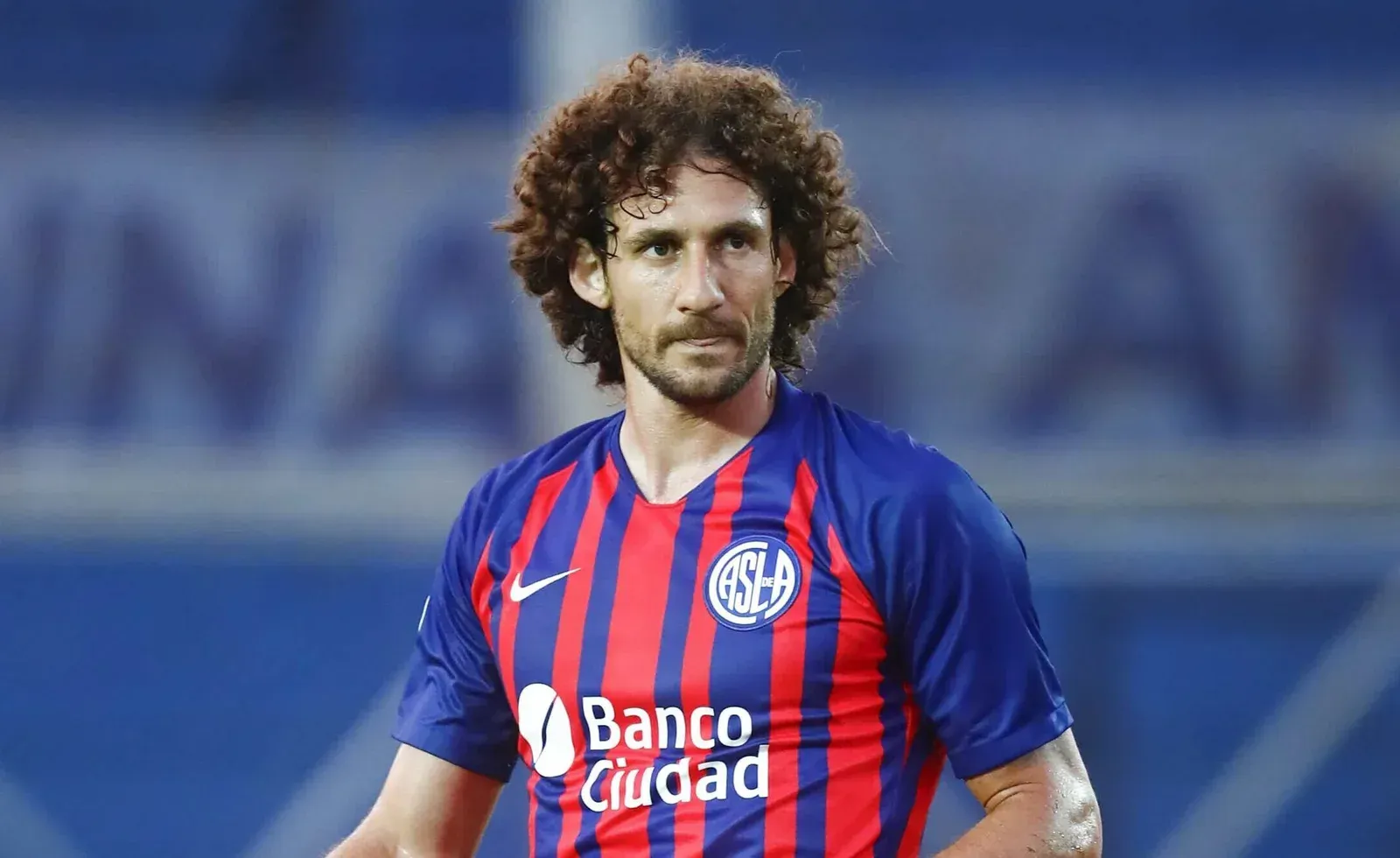 Jugador de fútbol con cabello castaño rizado con una camiseta de rayas azules y rojas, al aire libre.