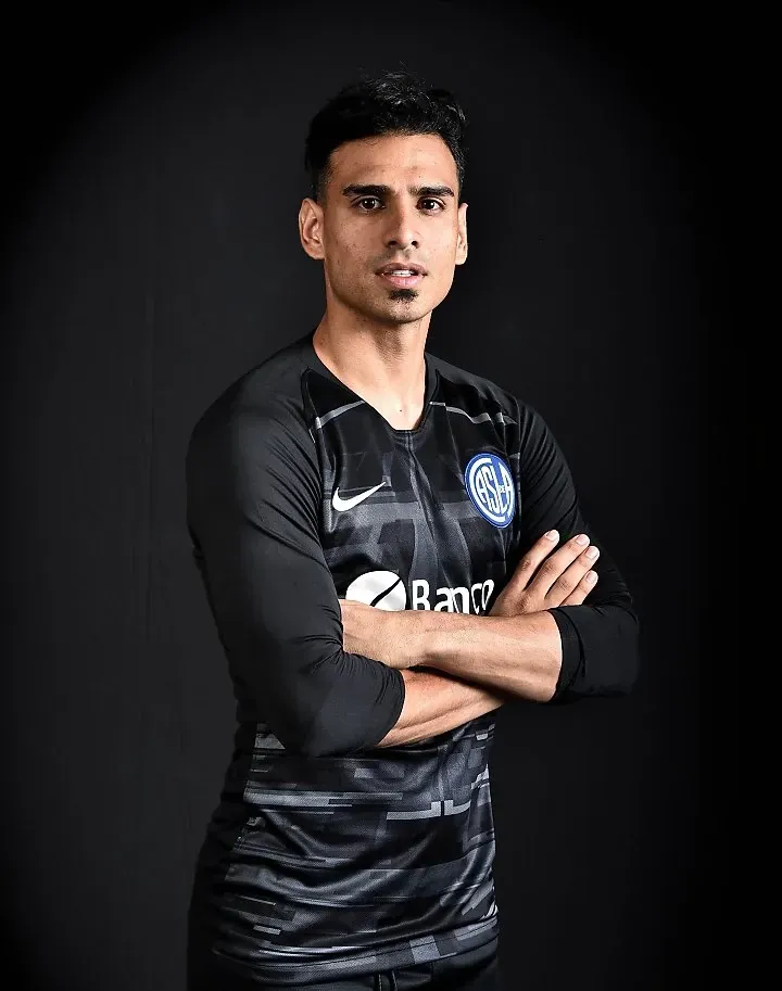 Jugador de fútbol con los brazos cruzados, vistiendo una camiseta negra y gris con patrocinadores sobre un fondo oscuro.