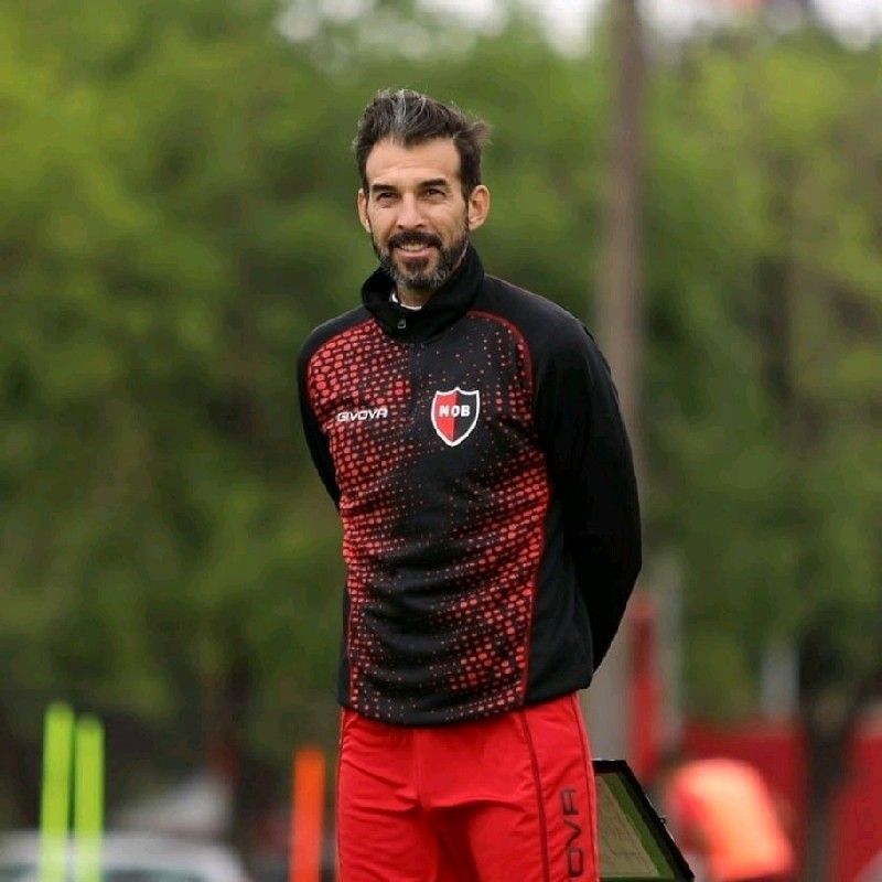 Un hombre con chaqueta deportiva y pantalones negros y rojos se encuentra en un campo con las manos detrás de la espalda.