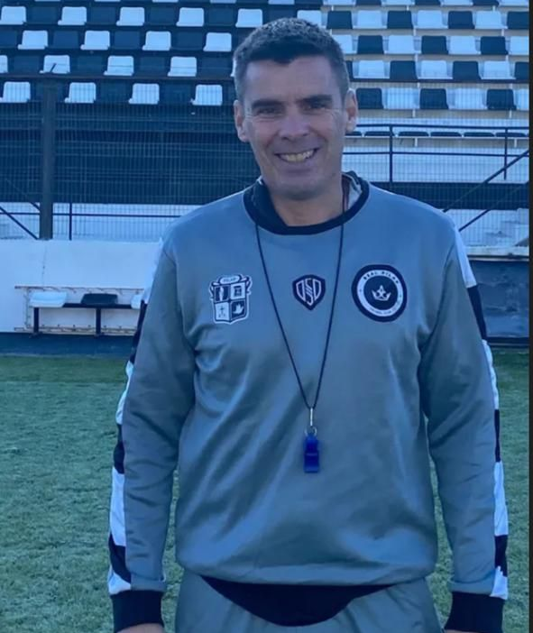 Hombre vestido con chándal gris, sonriente, con silbato, en el campo de fútbol, con el logo en el pecho.