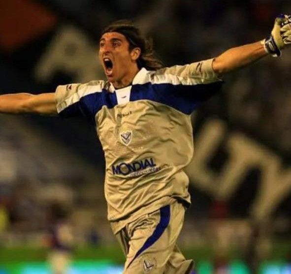 Portero con camiseta color canela y azul celebrando, con los brazos en alto y la boca abierta, al aire libre.