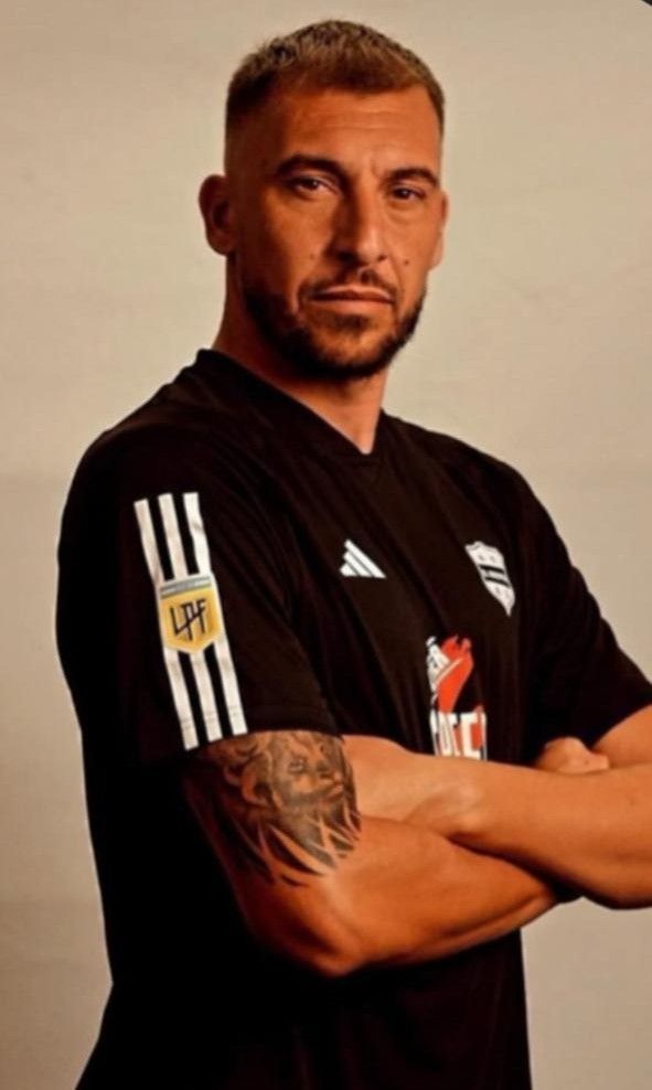 Hombre con camiseta Adidas negra, con los brazos cruzados, un tatuaje en la manga y una expresión severa.