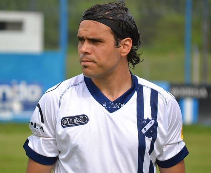 Jugador de fútbol con camiseta azul con los brazos cruzados sobre un fondo morado.