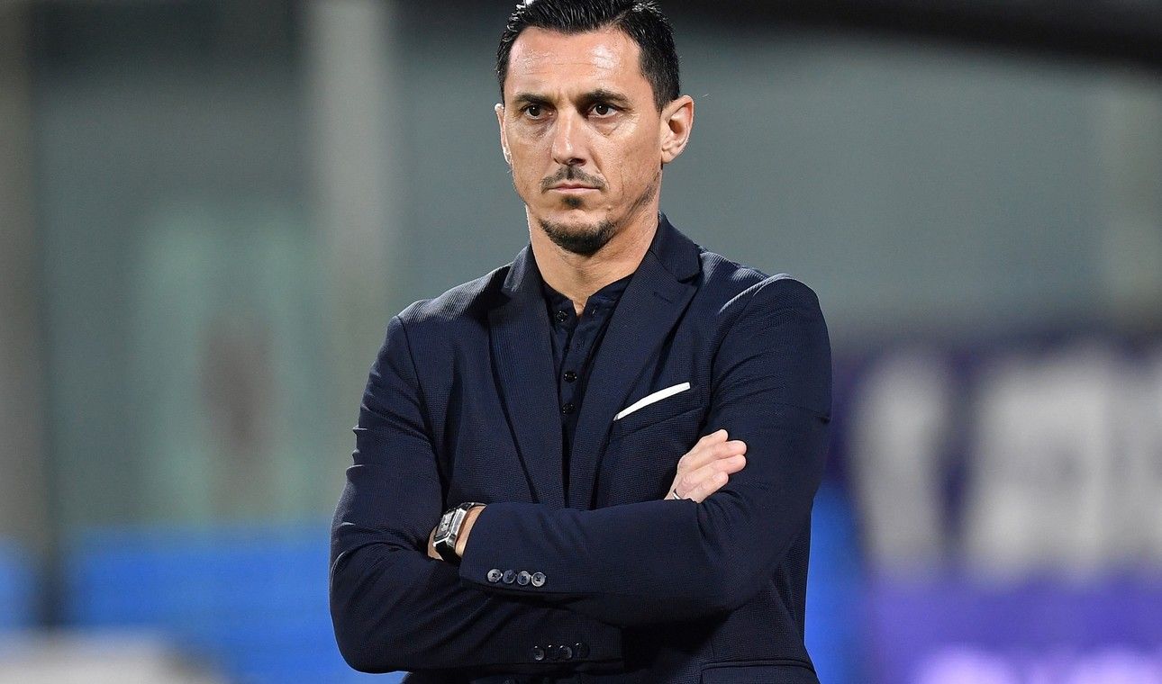 Entrenador de fútbol de pie, con los brazos cruzados, vestido con un traje oscuro y con aspecto serio.